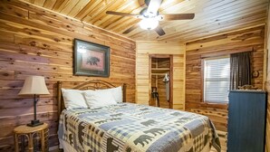 A wooden cabin bedroom with a bed, a nightstand, a ceiling fan, and a framed picture on the wall. - Papa Bear Log Cabin at the Buffalo River (Compton)