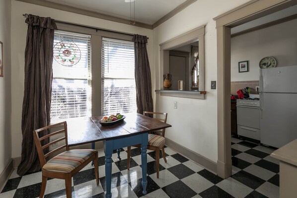 Un comedor con suelo de cuadros, una mesa de madera y sillas. Hay una ventana con cortinas, un florero y un reloj en la pared.