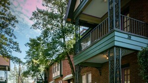 Une entrée fermée par un portail mène à un immeuble doté d’un balcon, entouré de plantes en pot et caractérisé par une façade en briques.