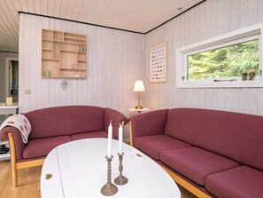 A room with a red sofa, a white table with candles, a lamp, a window with a view of greenery, and a wooden shelf with decorative items. - 5 Person Holiday Home in Tranekaer (Tranekaer)