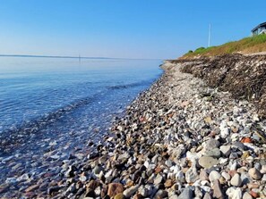 A pebbled beach with a clear view of the water and a distant bridge.