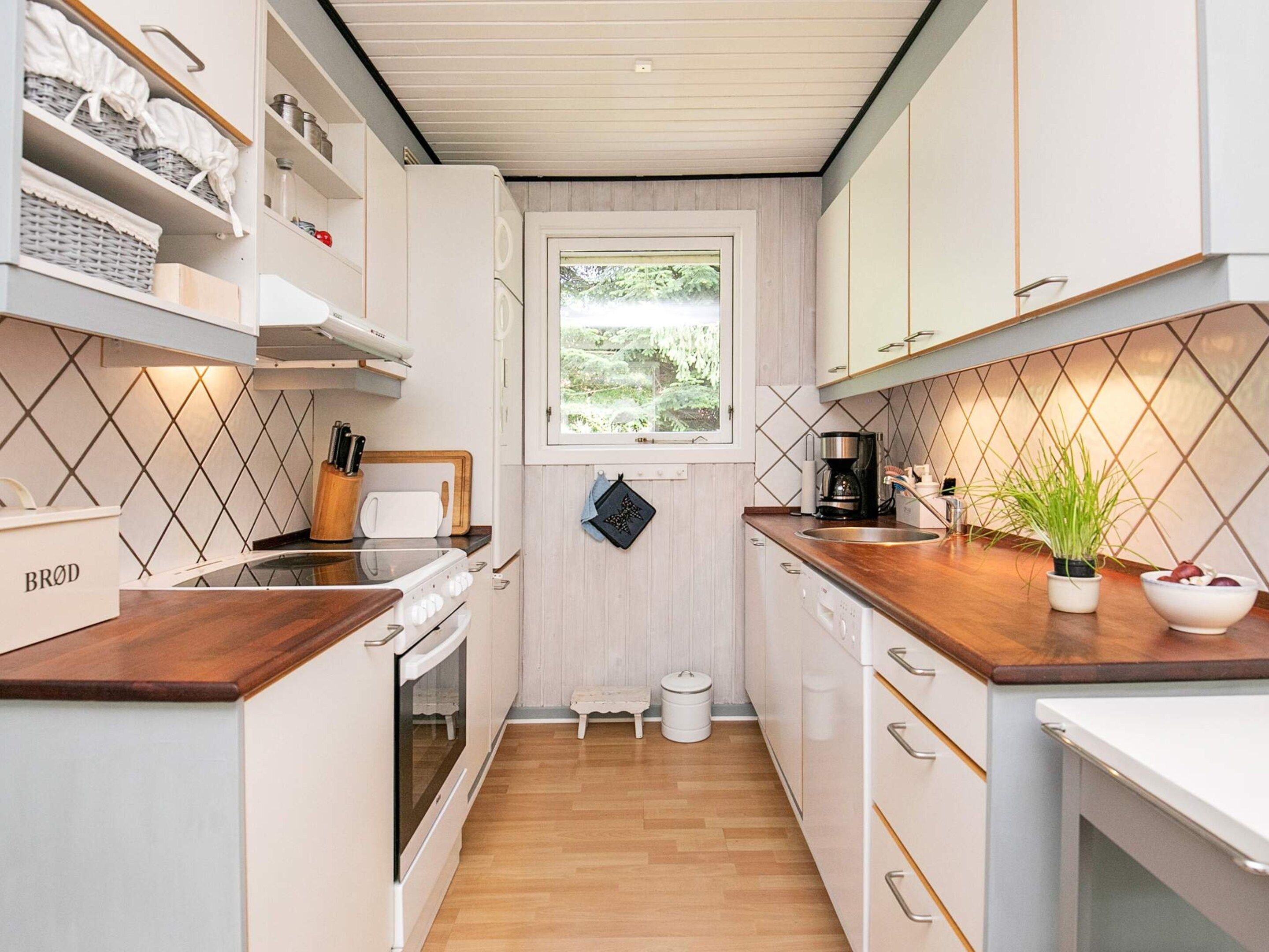 Cocina funcional con armarios blancos, encimera de madera, horno integrado y ventana con vistas a la vegetación.
