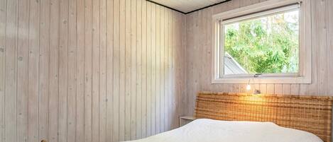 A room with a bed, wooden walls, a window with a view of greenery, and a ceiling light fixture.