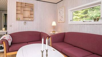 A room with a large red sofa, a white table with candles, a wooden shelf, and a window with a view of greenery.