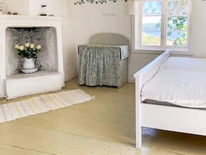 A bedroom with a white bed, a small table with a patterned tablecloth, a vase with flowers on a shelf, and a window with a view of greenery.
