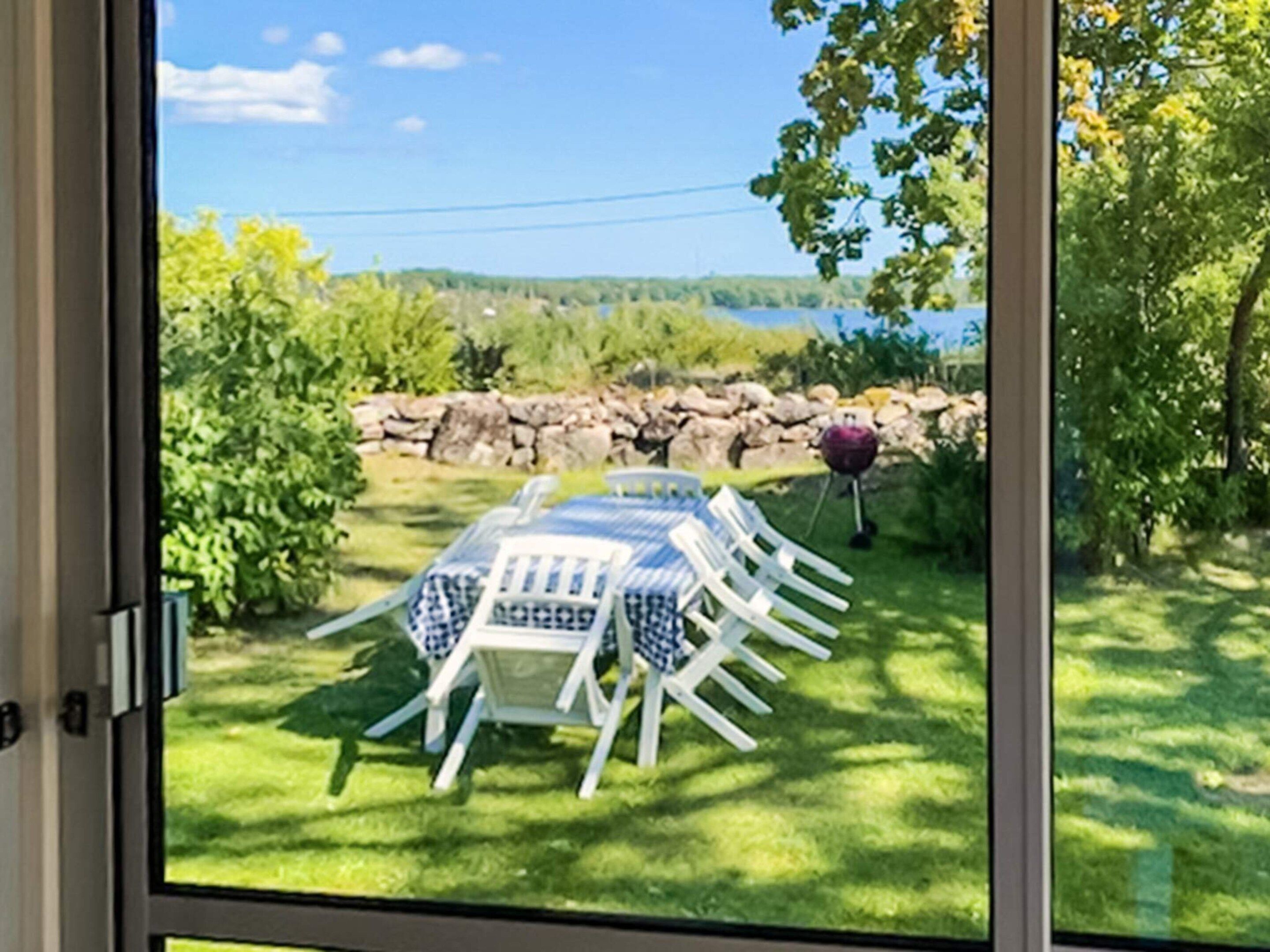 Vista pela janela de uma área de refeições com mesa e cadeiras, parede de pedra e vista para um lago e vegetação.
