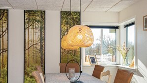 A dining area with a long table, wicker chairs, a woven pendant light, and a view of trees through a window.