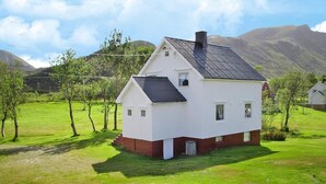 Ein weißes Haus mit dunklem Dach, umgeben von grünem Gras und Bäumen, mit Bergen im Hintergrund.