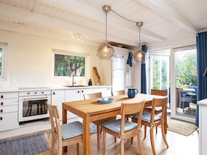 Een moderne keuken met witte kasten, een houten eettafel en stoelen. Er hangen hanglampen boven de tafel en er is uitzicht op het groen door een groot raam.