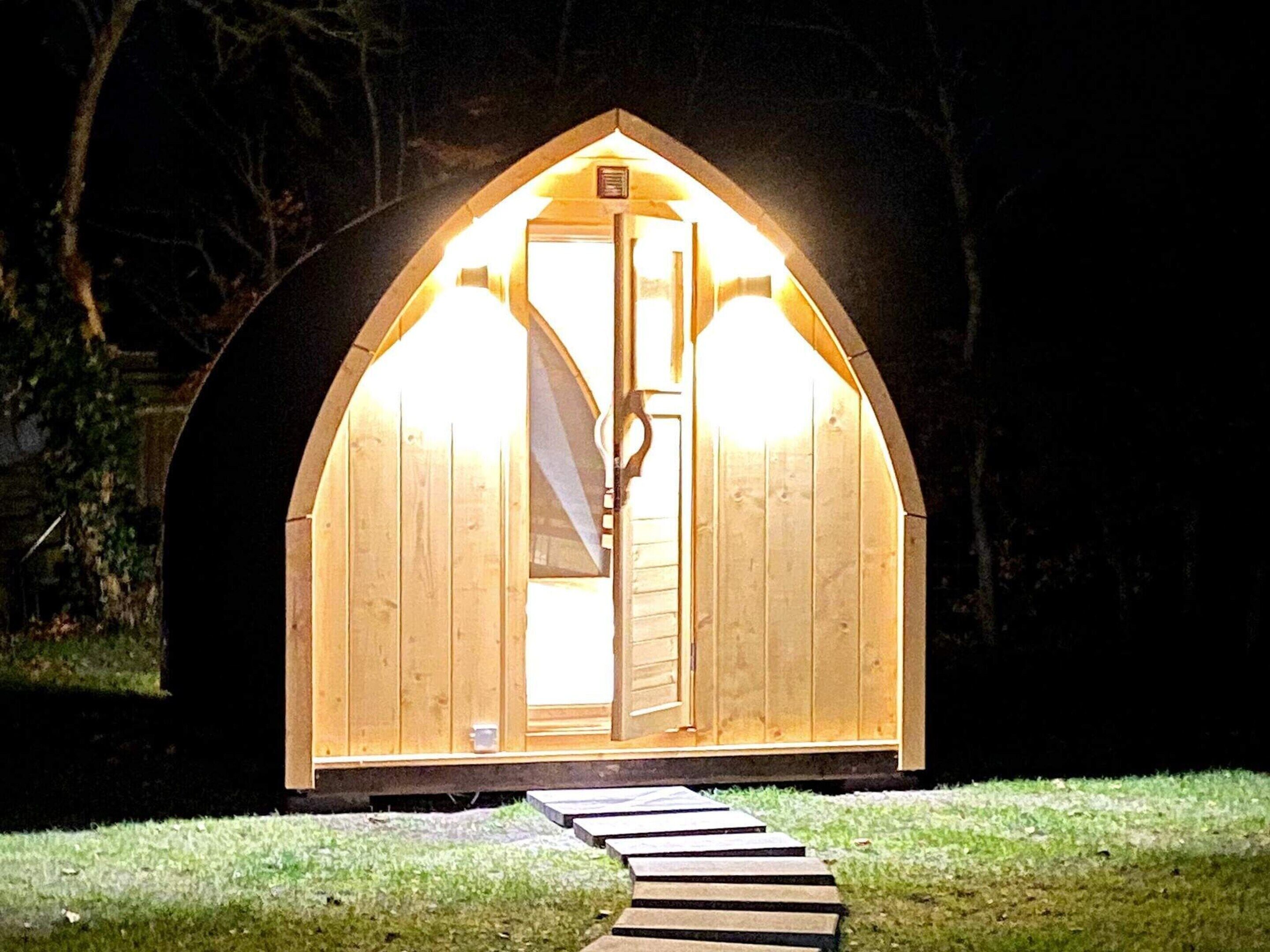 Una estructura de madera con techo a dos aguas e ingreso central iluminado desde el interior.