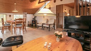 Un salon chaleureux avec un plafond en bois, une cheminée, un coin repas avec des chaises, un canapé, un téléviseur et une table en bois agrémentée d’un vase et de bougies.