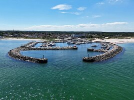Una marina circular con barcos amarrados, rodeada por una barrera rocosa y una playa con edificios al fondo.
