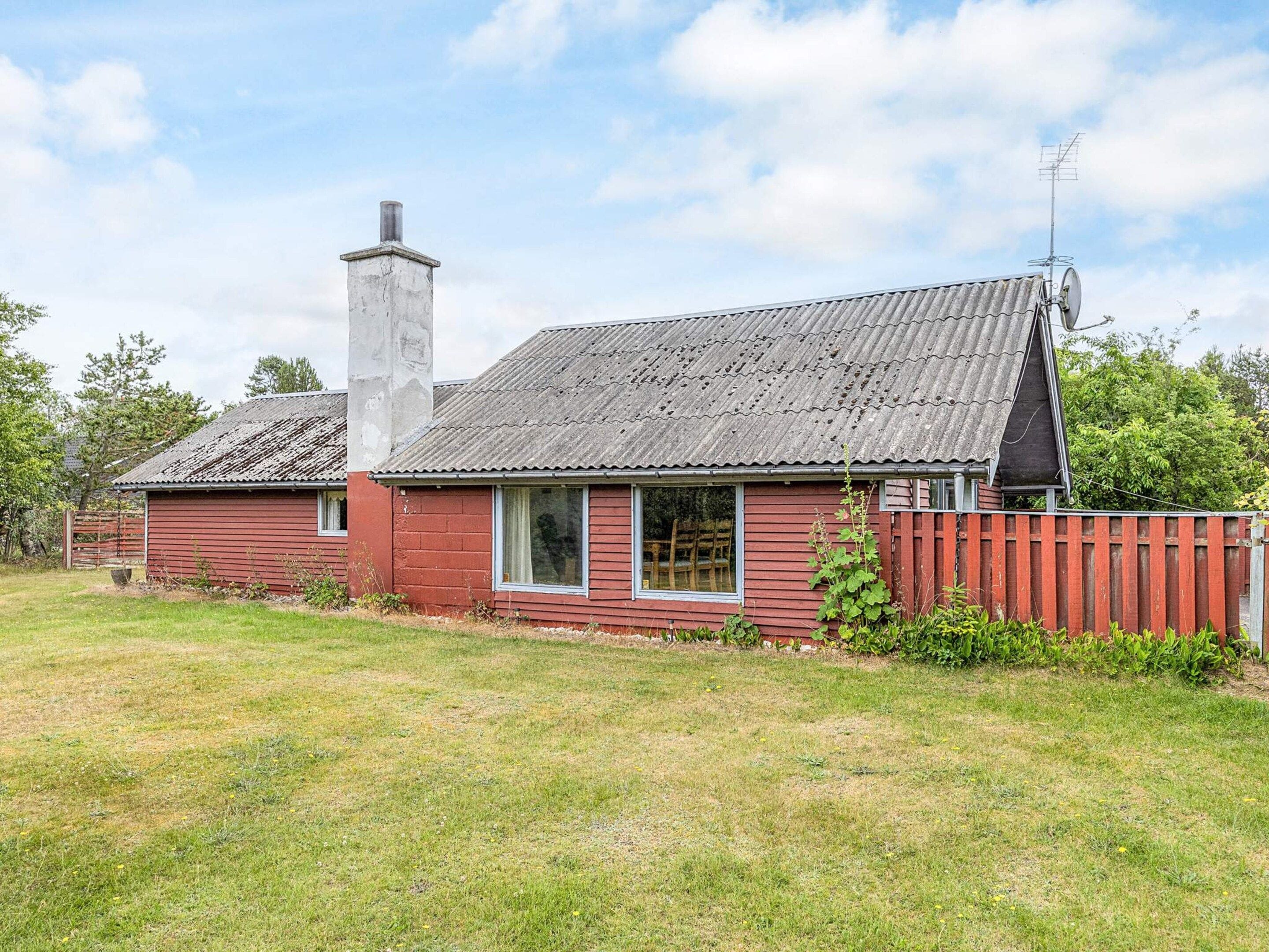 A red wooden house with a chimney, a fenced yard, and a metal roof.