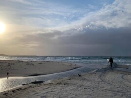 Una playa con una persona caminando por la orilla, olas rompiendo sobre la arena y un cielo nublado.
