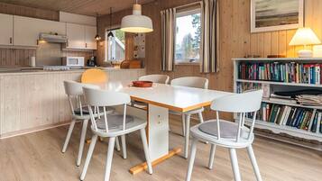 Una cocina moderna con armarios blancos y techo de madera, un comedor con mesa y sillas blancas, y una estantería llena de libros.