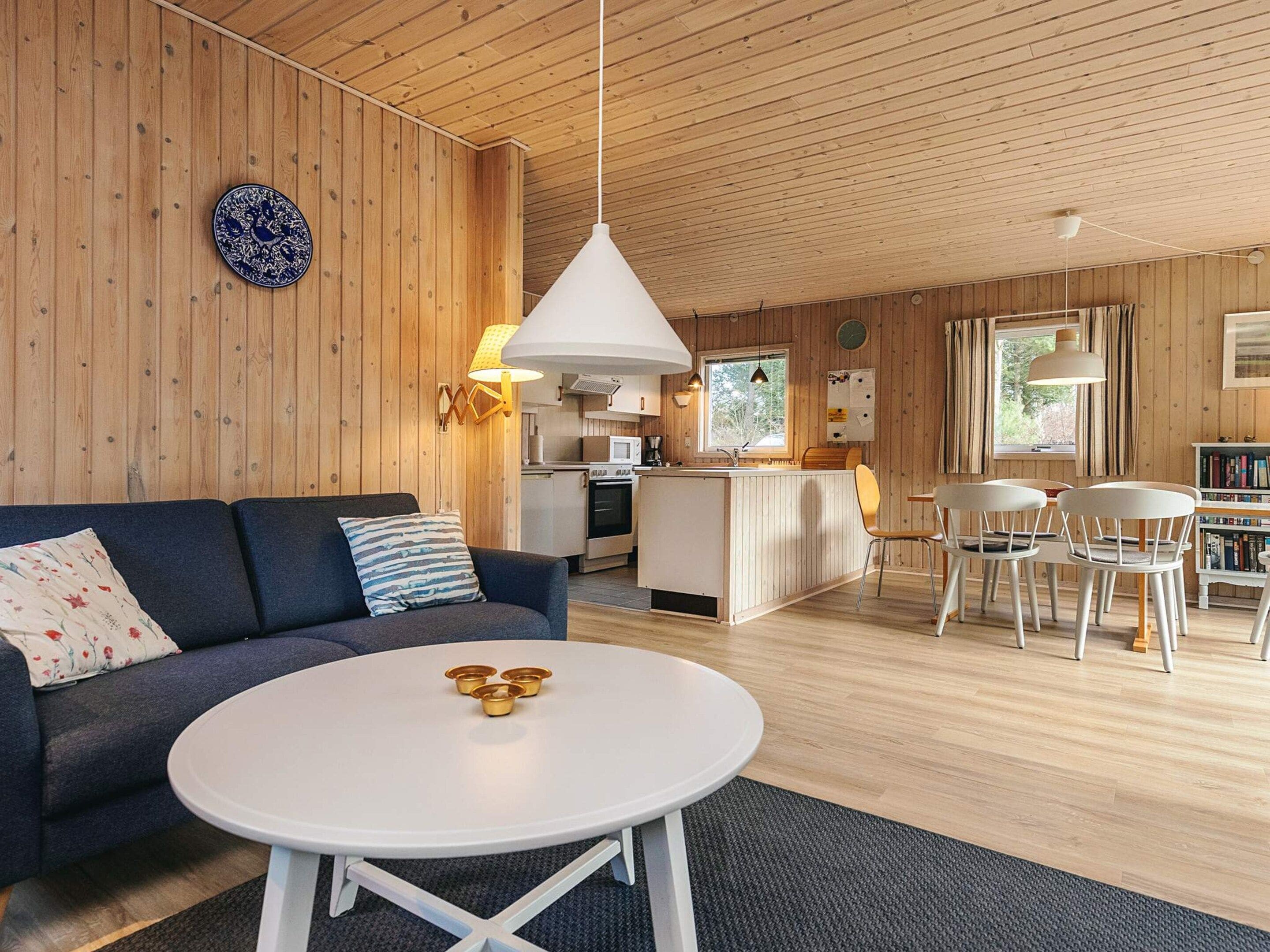 Un interior moderno con techo de madera, una mesa redonda blanca con cuencos dorados, un sofá azul oscuro con cojines estampados, y una zona de cocina con isla blanca.