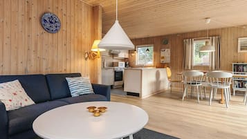 Un interior moderno con techo de madera, una mesa redonda blanca con cuencos dorados, un sofá azul oscuro con cojines estampados, y una zona de cocina con isla blanca.