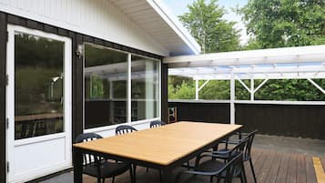 Un patio exterior con una mesa y sillas de madera, un vidrio que da a una zona cubierta y vistas a árboles.