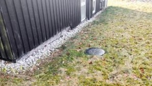 Fachada de un edificio con revestimiento metálico, cimientos de concreto y una zona verde con una rejilla de metal.