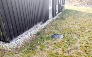 En byggnadsfasad med metallbeklädnad, en grund av betong och ett grönområde med ett metallgaller.