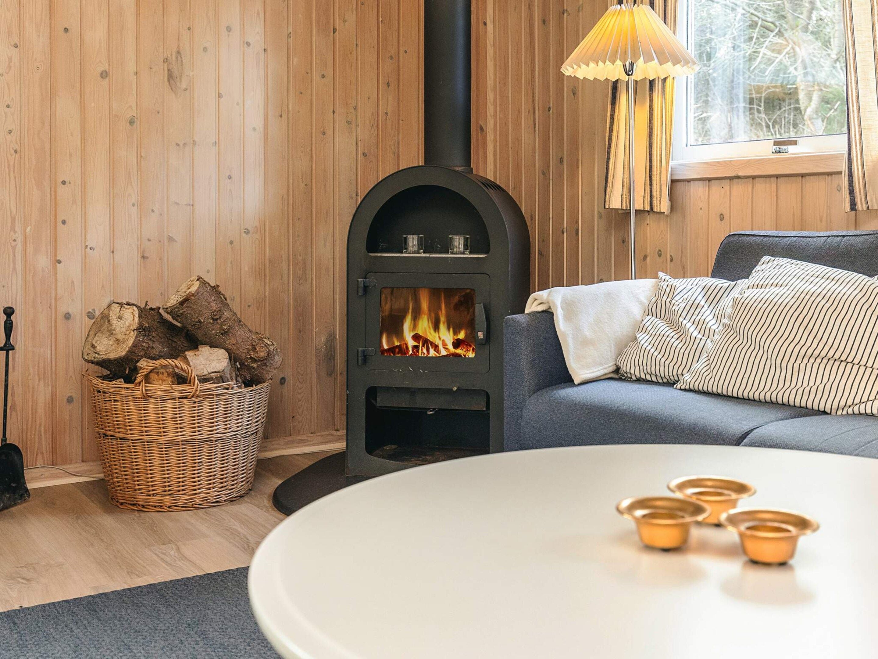 Une pièce chaleureuse avec un poêle à bois, un panier en osier rempli de bûches, un canapé gris et une table agrémentée de bougeoirs.