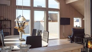 Ein gemütliches Zimmer mit großem Fenster, Kamin, Fernseher und einem Essbereich mit Stühlen und Tisch.