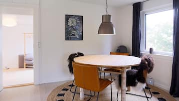 A dining area with a round table, chairs, a hanging light fixture, a painting on the wall, and a window with curtains.