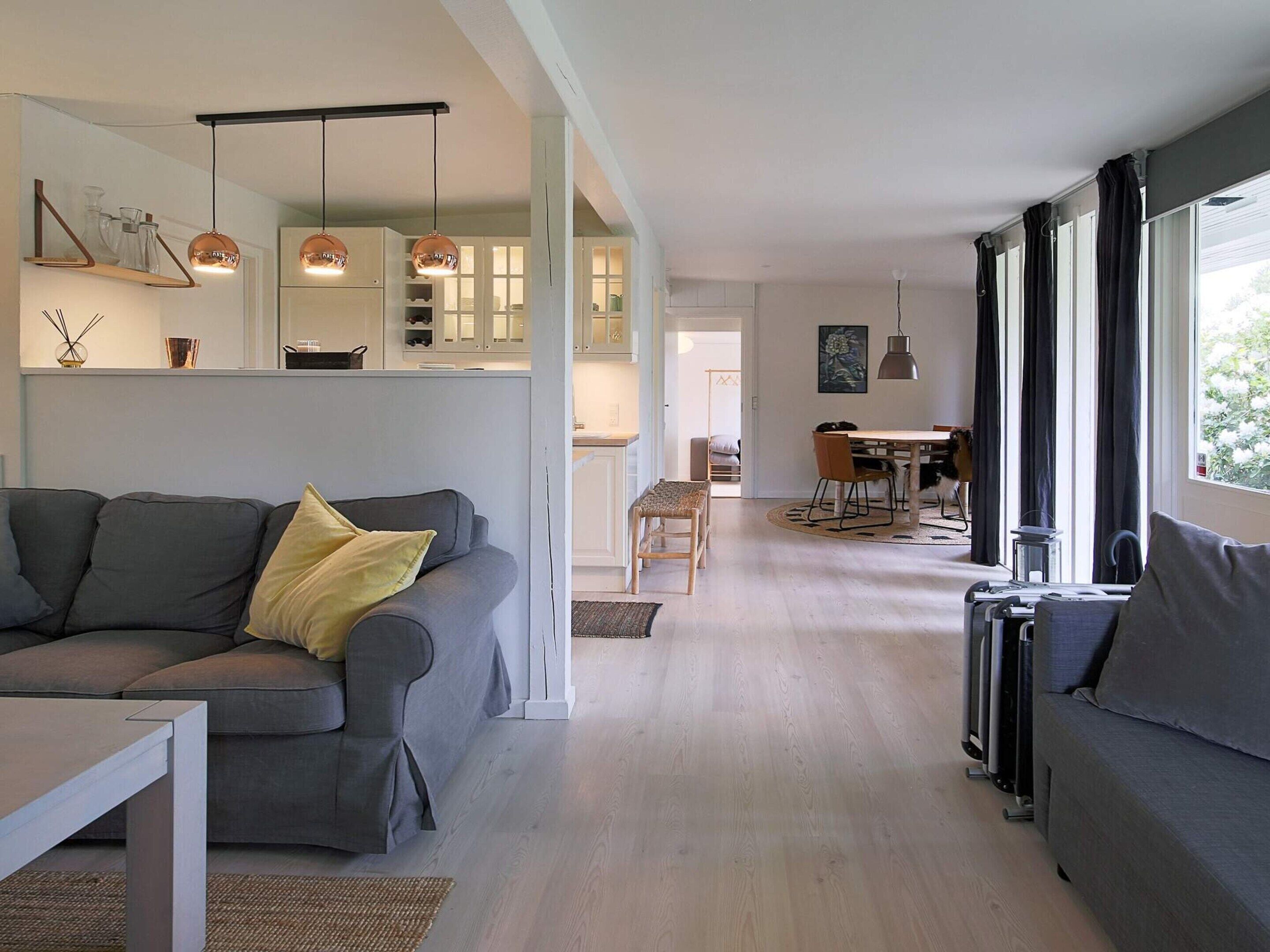 A modern living room with a grey sofa, a dining area with a table and chairs, and a kitchen with white cabinets and pendant lighting.