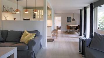 A modern living room with a grey sofa, a dining area with a table and chairs, and a kitchen with white cabinets and pendant lighting.