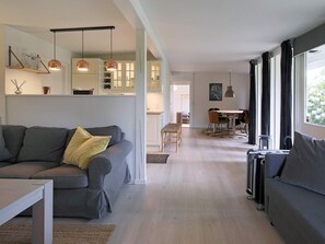 A modern living room with a grey sofa, a dining area with a table and chairs, and a kitchen with white cabinets and pendant lighting. - 4 Person Holiday Home in Gilleleje (Gilleleje)