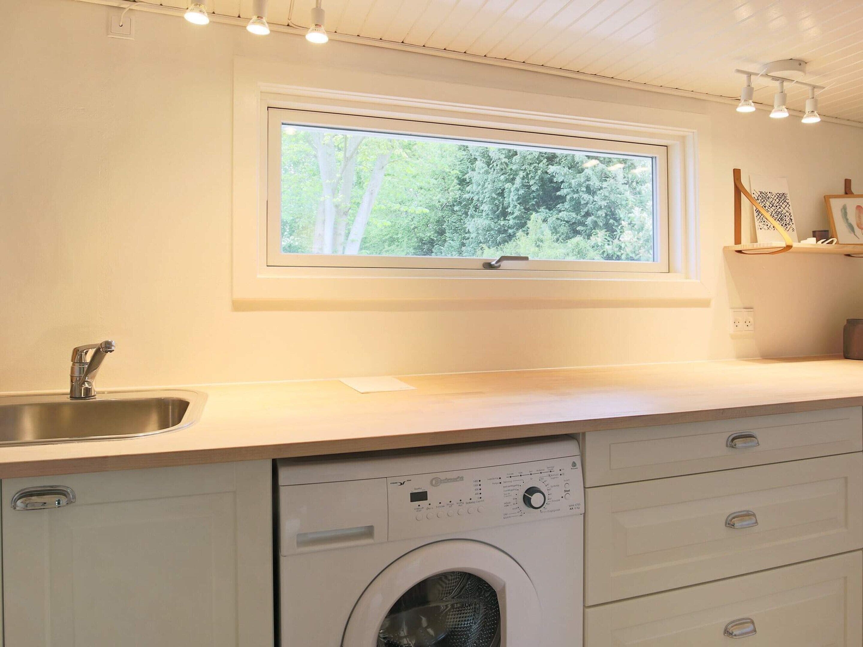 A kitchen with a built-in washing machine, a window with a view of trees, and a shelf with items.