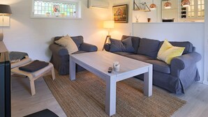 A modern living room with a grey sofa, a white coffee table, and a small wooden bench. There is a window with a stained glass design, a wall-mounted air conditioner, and a painting above the sofa. - 4 Person Holiday Home in Gilleleje (Gilleleje)