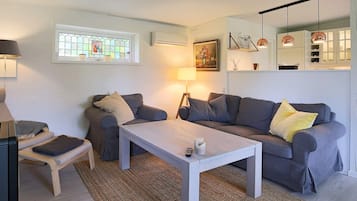 A modern living room with a grey sofa, a white coffee table, and a small wooden bench. There is a window with a stained glass design, a wall-mounted air conditioner, and a painting above the sofa.