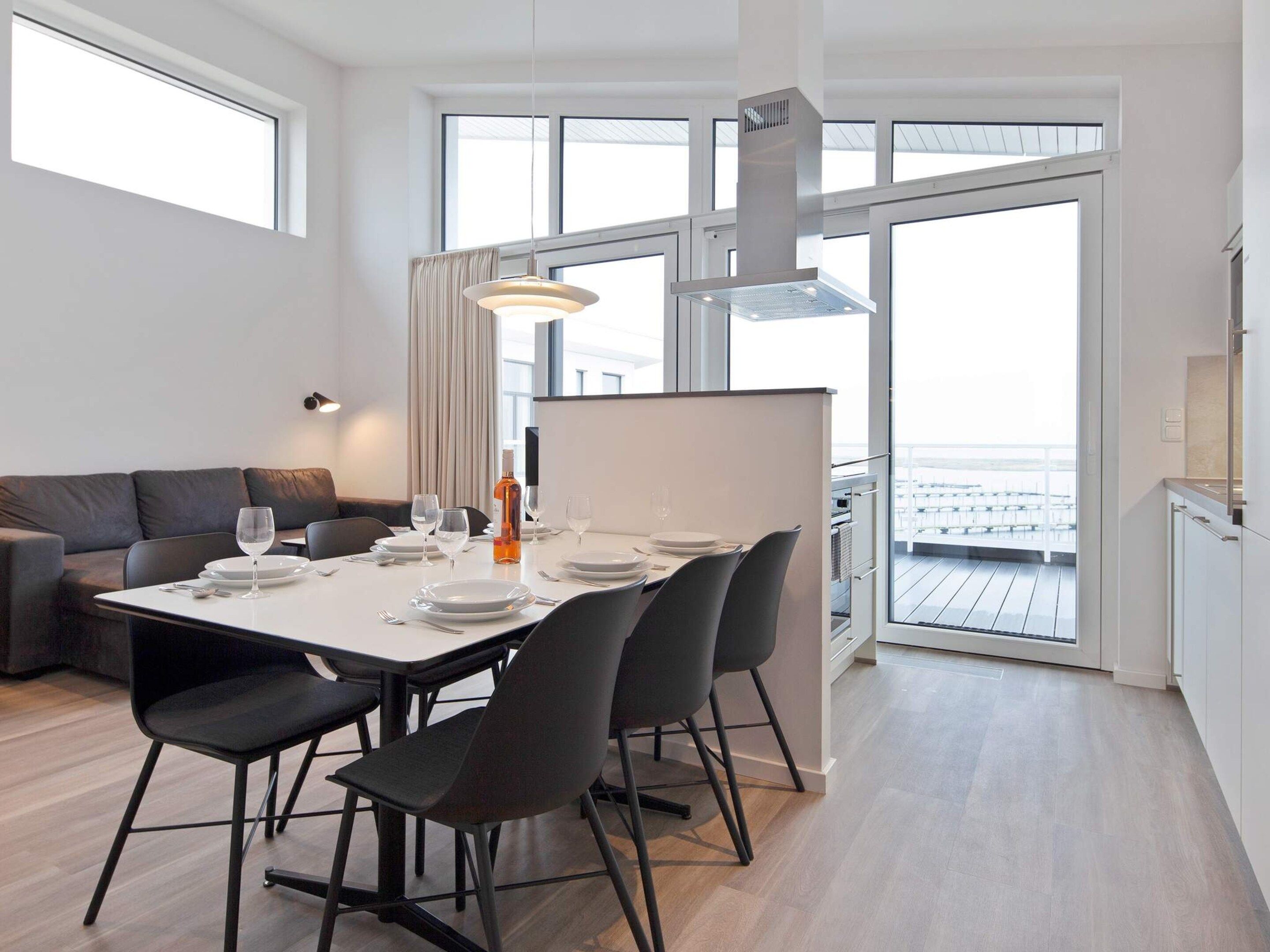 A modern dining area with a white table set for four, black chairs, a pendant light, and a view of the sea through large windows.