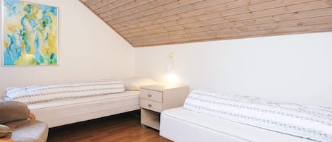 A cozy bedroom with a sloped ceiling, wooden floor, two beds with white bedding, a chair, and a painting on the wall.