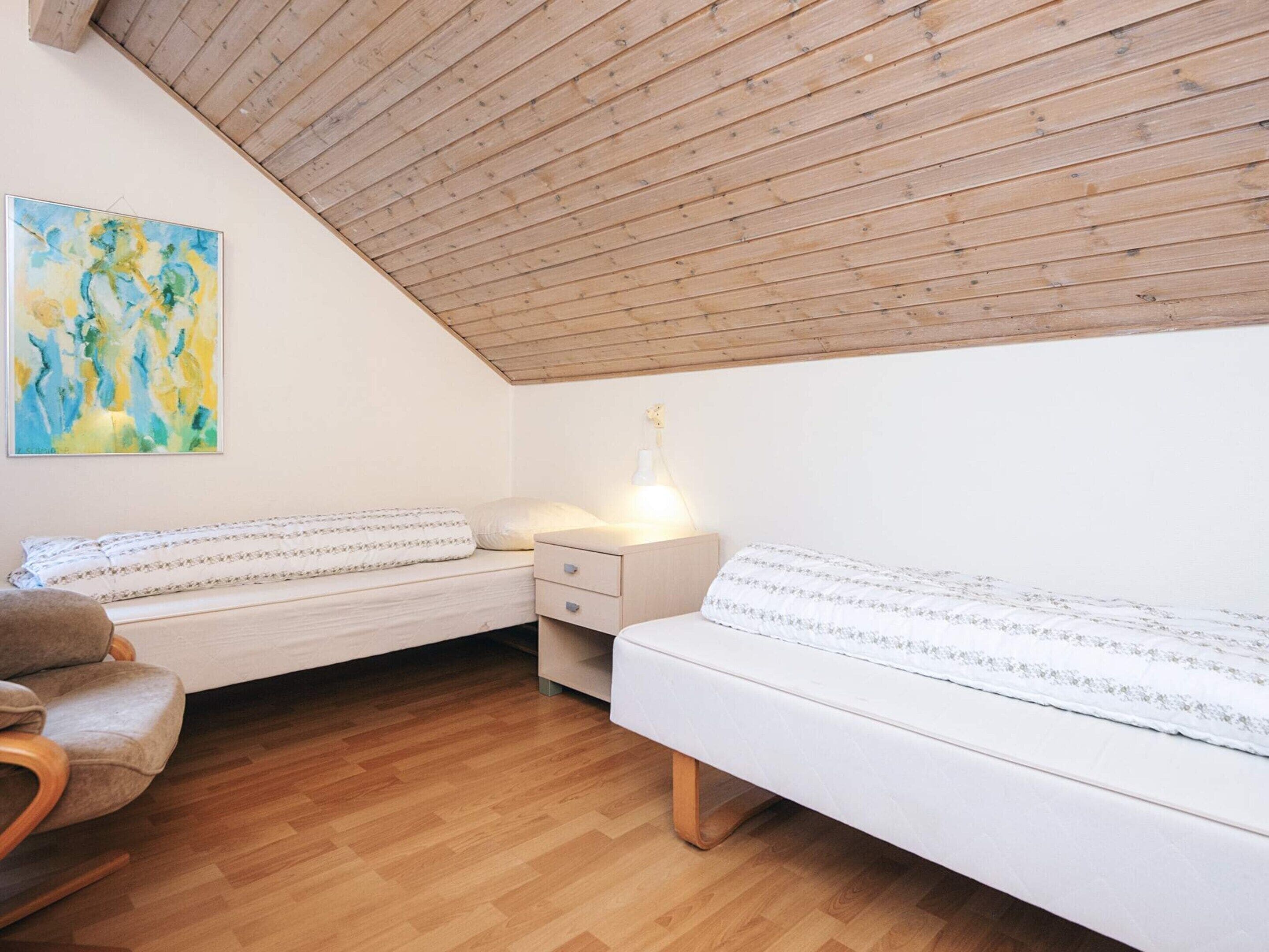 Ein gemütliches Schlafzimmer mit schrägen Wänden, Holzboden, zwei Betten mit weißer Bettwäsche, einem Sessel und einem Bild an der Wand.