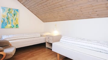 Ein gemütliches Schlafzimmer mit schrägen Wänden, Holzboden, zwei Betten mit weißer Bettwäsche, einem Sessel und einem Bild an der Wand.