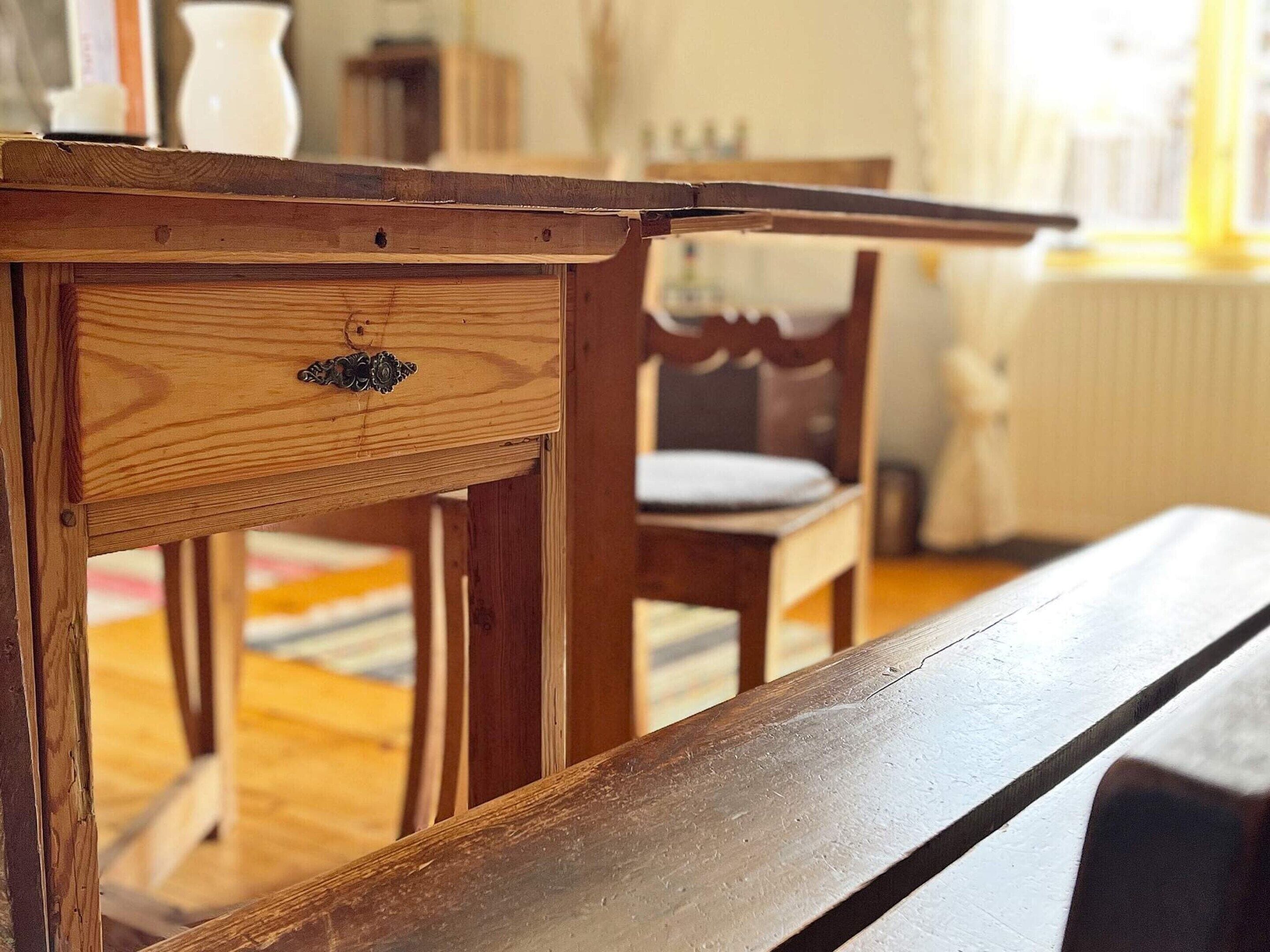 A wooden table with a drawer and a bench in a room with a chair and a radiator in the background.