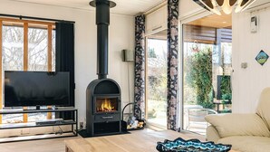 A modern living room with a wood-burning stove, a flat-screen TV, and a large window offering a view of the outdoors. - 6 Person Holiday Home in Laeso (Læsø)