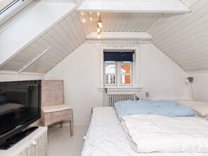 Ein kleines, weißes Schlafzimmer im Dachgeschoss mit Fernseher, Stuhl, einem Bett mit Kissen, einem Fenster mit Blick auf ein Haus und einer Heizung.