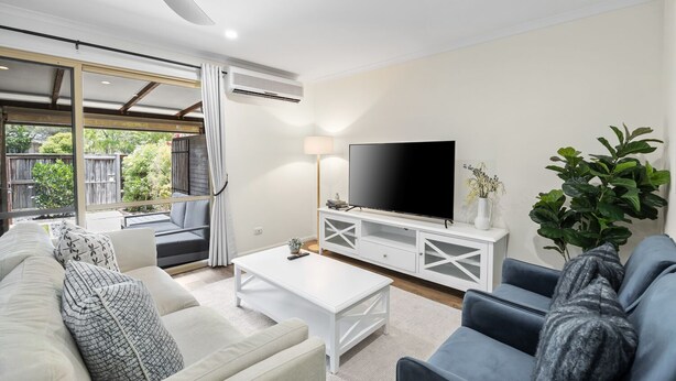 A modern living room with a flat-screen TV, white furniture, and a large window overlooking a backyard. - Regency 3 Bedroom Central Town House (Sunshine Coast)