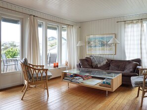 Un salon avec un canapé en cuir marron, une table basse en verre sur laquelle sont posés des livres, un plancher en bois et une vue sur l’extérieur grâce à de grandes fenêtres.