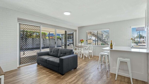 A modern living room with a grey sofa, a dining area with white chairs, and a kitchen with a white countertop and bar stools.