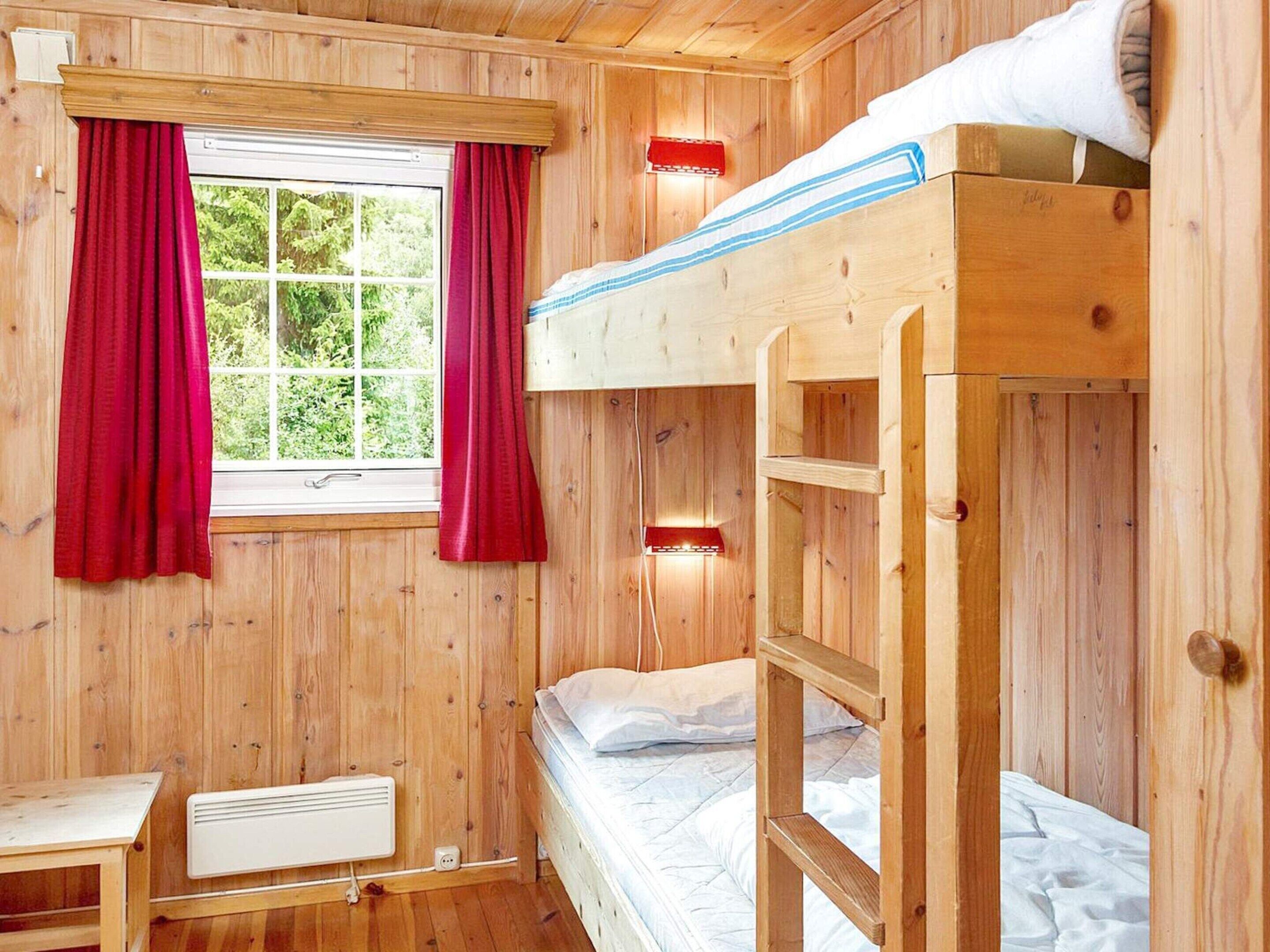 A wooden cabin room with a bunk bed, a window with red curtains, a heater, and a small table.