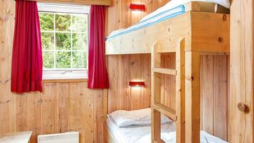 A wooden cabin room with a bunk bed, a window with red curtains, a heater, and a small table.