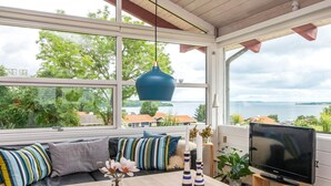 A modern living room with a sofa, a coffee table, and a TV on a wooden stand. Large windows offer a view of trees and a body of water. - 6 Person Holiday Home in Aabenraa-by Traum (Aabenraa)