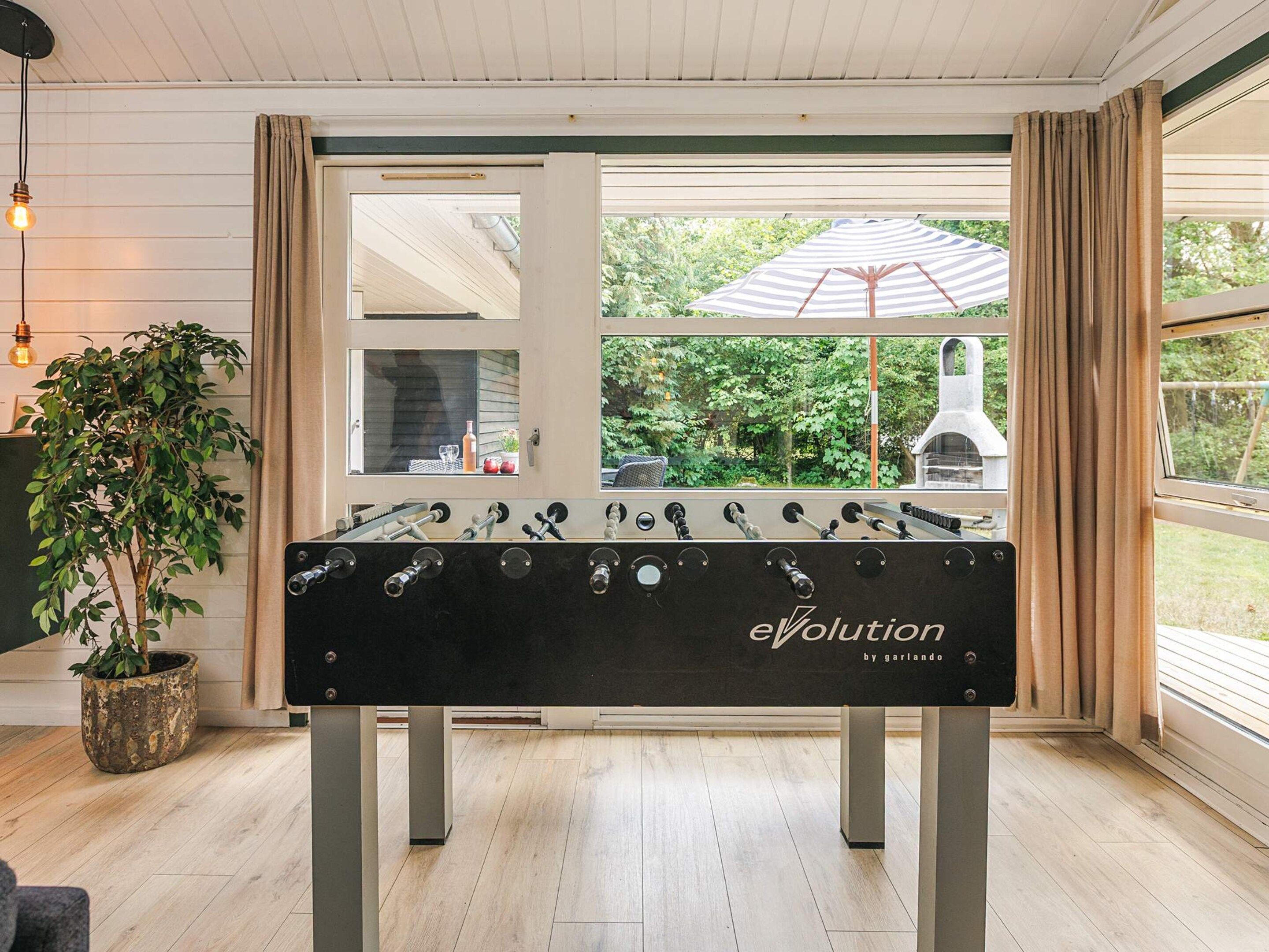 A foosball table with the brand 'evolution' in a room with wooden flooring, a potted plant, and a view of greenery outside.