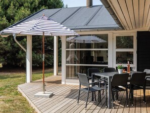 Eine überdachte Terrasse mit Tisch und Stühlen, einem gestreiften Sonnenschirm und Blick auf das Haus durch große Fenster.