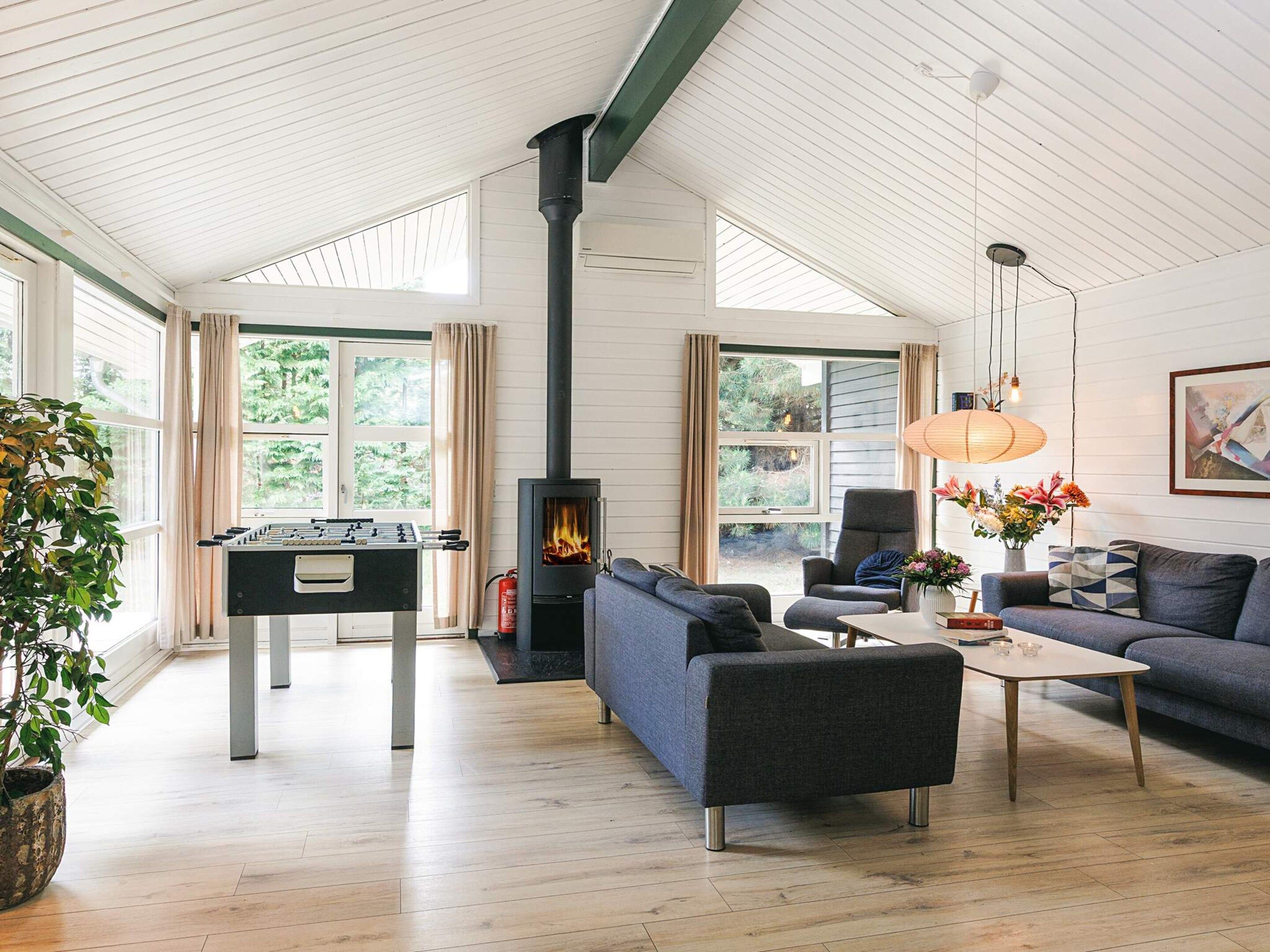 A spacious living room with a sofa, a coffee table, and a foosball table. There is a fireplace with a chimney, and the room has large windows.