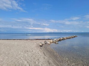 A beach with a rocky breakwater extending into calm waters, a clear sky, and a distant shoreline.